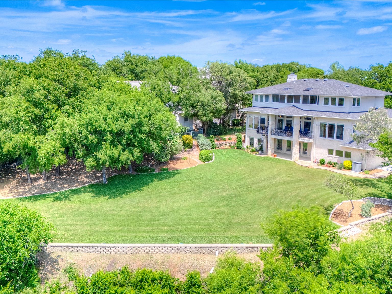 Rear elevation of the estate viewed from the back lawn: three levels with a covered porch on the lower level and a balcony on the upper level, large windows facing the lawn, mature oaks framing the property.