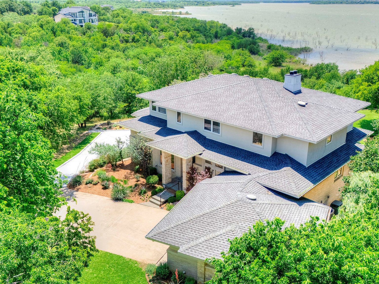 Overhead view of the home's roofline with Joe Pool Lake spread across the upper third of the frame: gray composite roof, multiple chimneys, motor court visible to the left, dense tree canopy ringing the structure.