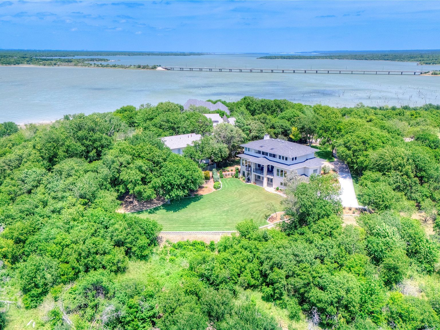 Wide aerial showing the estate centered in a clearing of mature trees with Joe Pool Lake wrapping around the peninsula on three sides, the lake bridge visible in the distance.